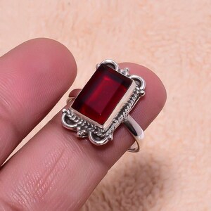 Könnte beinhalten: Ein Silberring mit einem rechteckigen, tiefroten Edelstein. Der Edelstein ist in einer verzierten Silberfassung mit strukturiertem Design gefasst. Der Ring wird auf einem Finger gezeigt.