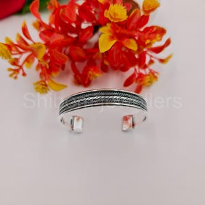 May include: A silver cuff bracelet featuring a central band with a detailed braided rope texture flanked by smooth polished edges. The metal surface displays a high-shine finish with darkened recessed areas that highlight the intricate woven pattern.