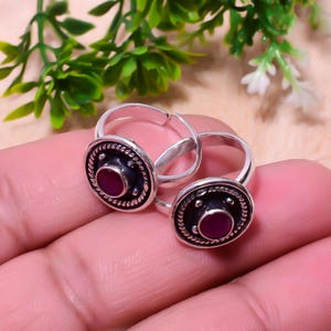 May include: Two silver toe rings, each featuring a dark, textured border and a central, round, red gemstone. The rings are set against a blurred backdrop of green leaves and skin.
