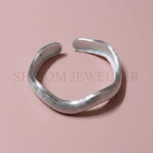 Könnte beinhalten: Offener, silberfarbener Ring mit strukturiertem, organischem Design. Der Ring hat ein mattes Finish und eine leicht unregelmäßige Form. Der Hintergrund ist einfarbiges, gedämpftes Rosa. Der Text "SHOOM JEWELER" ist sichtbar.