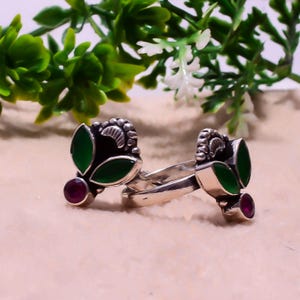 May include: Two silver rings with leaf and berry designs. Each ring has a dark green leaf-shaped stone, a small round red stone, and a decorative silver top. The rings are set against a soft, neutral background.