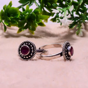 May include: Two silver rings, each featuring a circular, textured setting and a central, deep red gemstone. The rings are displayed against a soft, neutral backdrop, with green and white foliage in the upper corners.