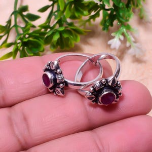 May include: Two silver rings, each featuring a floral design and a central, round, red gemstone. The rings are interlinked. The floral detailing has small, dark accents. Green foliage is visible in the background.