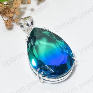 Puede incluir: Un colgante en forma de pera con una piedra preciosa facetada que presenta una transición de color degradado desde el azul profundo en la base hasta un verde azulado vibrante en la parte superior, engastado en un marco de metal plateado.