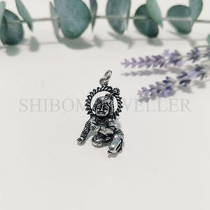 Könnte beinhalten: Silberanhänger mit detaillierter Darstellung einer sitzenden Gottheit mit Heiligenschein. Der Anhänger weist filigrane Details auf und ist an einer kleinen Schlaufe aufgehängt. Das Wort "SHIBONVELLER" ist im Hintergrund aufgedruckt.