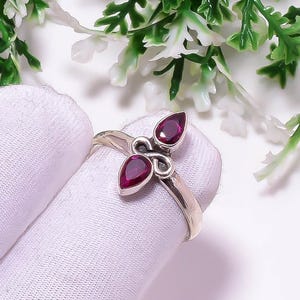 Puede incluir: Un anillo de plata con tres piedras preciosas rojas oscuras en forma de lágrima. El anillo presenta un diseño central en espiral y una banda dividida. Las piedras están engarzadas con garras, y se muestra sobre una superficie blanca.