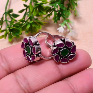 May include: Two silver rings with a floral design. Each ring features a central green stone surrounded by maroon petal-shaped stones. The rings are displayed against a soft, neutral background with green foliage.