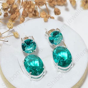 May include: A pair of drop earrings featuring two faceted teal-colored stones set in silver-toned metal. Each earring consists of a smaller round stone positioned above a larger oval stone, both secured with four-prong settings on a white display.