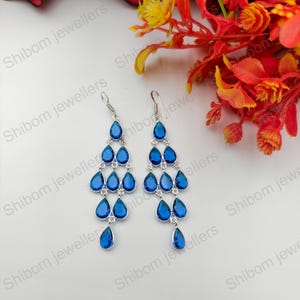 Puede incluir: Un par de pendientes tipo candelabro en tono plateado con múltiples piedras azules en forma de lágrima dispuestas en un diseño en cascada. Cada pendiente cuelga de un gancho simple, con las piedras azules unidas por pequeños anillos de metal.
