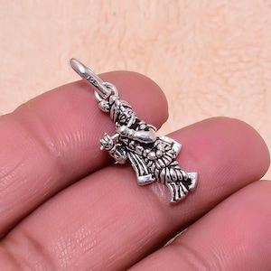 Puede incluir: Un pequeño y detallado colgante de plata con forma de figura, posiblemente una deidad maya o azteca. El colgante tiene intrincadas tallas y está suspendido de un pequeño lazo. La figura lleva un tocado y ropa adornada.