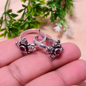 May include: A pair of silver toe rings with detailed, floral patterns. Each ring has a small, circular, deep red gemstone in the centre. The rings are shown against a soft, neutral backdrop, with green foliage visible in the top part of the image.