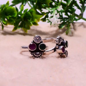 May include: Two silver rings with intricate detailing. Each ring is adorned with a clear, round gemstone, a deep red stone, and a green stone. The rings are displayed against a soft, neutral backdrop.