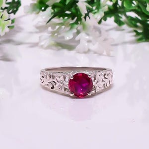 May include: A silver ring with an oval, deep red gemstone. The band features an intricate floral and scrollwork design. The ring is set against a white background with blurred greenery.