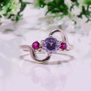 May include: A silver ring with a central, round, lilac gemstone and two smaller, magenta gemstones. The ring has a distinctive, flowing design with a heart-shaped element. The gemstones are set in a silver band.
