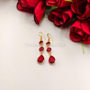 May include: A pair of gold-tone drop earrings featuring two circular red stones and one teardrop-shaped red stone suspended from a hook wire. The text Shibori Jeweller is visible in the centre. The earrings are set against a background of red roses.