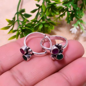 May include: Two silver toe rings with decorative settings. Each ring features a cluster of three gemstones: a central oval ruby and two smaller stones, one green and one red. The rings are displayed on a light pink surface.