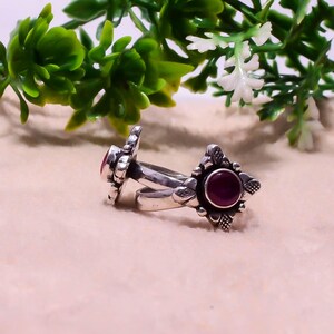 May include: Two silver rings with a floral design and a round, dark red gemstone. The rings are set against a neutral background with green and white foliage in the background. The rings are likely made of sterling silver.