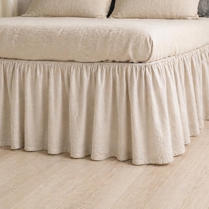 Op de afbeelding: Een bed met een beige linnen bedrok en bijpassend beddengoed. De bedrok heeft een gerimpeld ontwerp en het beddengoed bevat kussens. De kamer heeft een lichtgekleurde houten vloer.