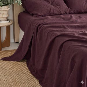 May include: A bed dressed with a deep burgundy linen sheet set. The sheets are draped over the bed, with a pillow visible. The bed is set on a woven rug. The sheets' colour is a rich, dark red-purple.