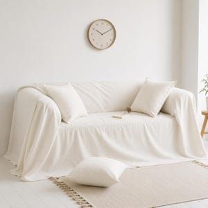 Op de afbeelding: Een crèmekleurige bank bedekt met een bijpassende stoffen hoes, aangevuld met drie vierkante kussens. Een ronde klok met een licht houten frame hangt aan de witte muur erboven. Een beige tapijt met franjes ligt op de vloer.