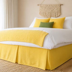 May include: A bed with white sheets, yellow pillows, and a yellow blanket. The bed skirt is also yellow. A green pillow and a woven wall hanging are also visible.