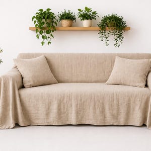 Op de afbeelding: Een beige linnen bank met twee bijpassende kussens en een gedrapeerde hoes. Een kleine houten bijzettafel heeft een vaas met bloemen. Groene planten in potten en hangend aan een plank voegen een vleugje natuur toe.