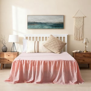 May include: A bedroom scene featuring a bed with a light pink bed skirt. The bed is made with white pillows and a beige patterned pillow. A painting hangs above the bed, and a macrame wall hanging is on the right.