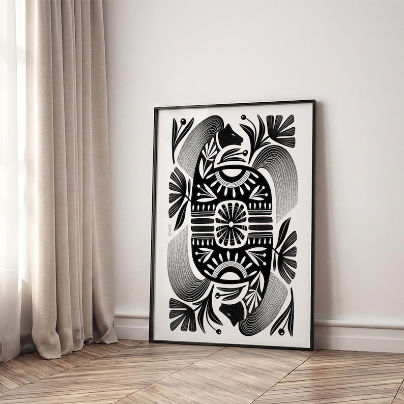 May include: Black and white framed art print featuring a symmetrical design. The artwork showcases a central geometric shape with floral and abstract elements. The print is displayed on a wooden floor, leaning against a white wall.