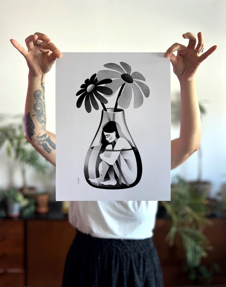 以下が含まれることがあります： ガラスの花瓶の中に女性と大きな花が2つ描かれた白黒のアートプリント。 女性は座って膝を抱えています。 花は大きな花びらを持っており、すべて白黒で表現されています。