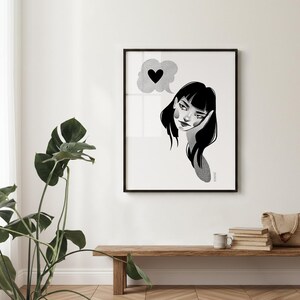 May include: Black and white framed art print featuring a stylized illustration of a woman with a thought bubble containing a heart. The artwork is displayed on a white wall above a wooden bench with a plant and other decorative items.