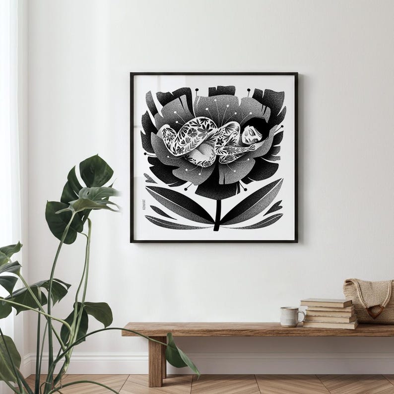 May include: Black and white art print in a black frame. The artwork depicts a person lying in a large flower. The person has detailed floral tattoos. The flower and person are in shades of gray. The print is displayed on a white wall.