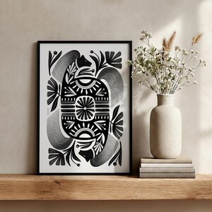 May include: Black and white framed art print with an abstract design. The artwork features symmetrical floral and geometric patterns. A vase with white flowers and a stack of books are on a wooden shelf next to the framed print.