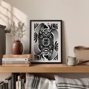 May include: Black and white framed art print with an abstract design. The artwork features symmetrical floral and geometric patterns. The print is displayed on a wooden shelf with a vase, books, and a mug.