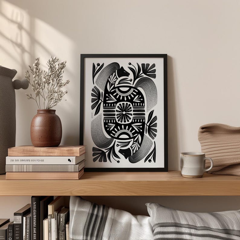 May include: Black and white framed art print with an abstract design. The artwork features symmetrical floral and geometric patterns. The print is displayed on a wooden shelf with a vase, books, and a mug.