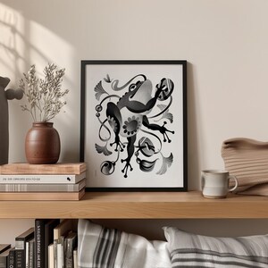 May include: Framed monochrome art print depicting a stylized frog with floral elements. The print is displayed on a wooden shelf alongside a vase, books, and a mug. The frog illustration is the central focus of the artwork.