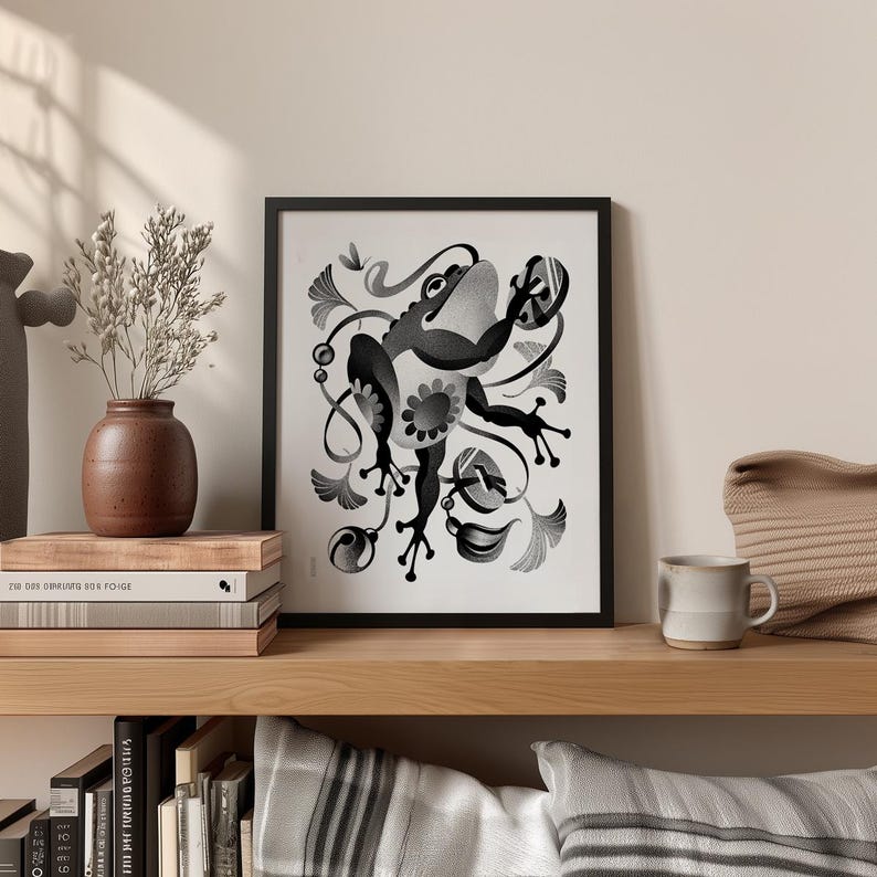 May include: Framed monochrome art print depicting a stylized frog with floral elements. The print is displayed on a wooden shelf alongside a vase, books, and a mug. The frog illustration is the central focus of the artwork.