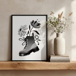 May include: Black and white framed art print featuring a combat boot with flowers. The illustration shows a gray and black boot with a thick sole, and two flowers emerging from the top. The artwork is displayed on a wooden shelf.
