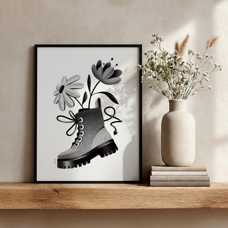 May include: Black and white framed art print featuring a combat boot with flowers. The illustration shows a gray and black boot with a thick sole, and two flowers emerging from the top. The artwork is displayed on a wooden shelf.