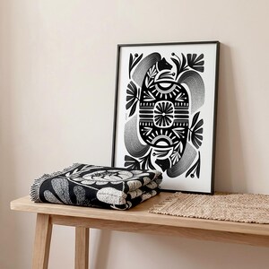 May include: A black and white floral throw blanket with fringe is folded on a wooden table. A framed black and white art print with a symmetrical design of floral and geometric shapes is propped up behind the blanket.