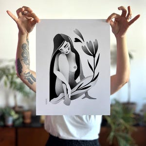 Może przedstawiać: Czarno-biały wydruk artystyczny przedstawiający siedzącą postać z długimi włosami, kwiatem i liśćmi. Grafika jest trzymana przez osobę. Postać siedzi ze skrzyżowanymi nogami. Kwiat i liście są stylizowane.