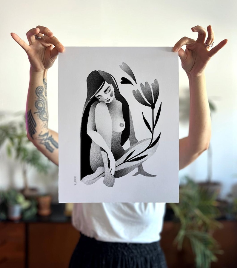 Może przedstawiać: Czarno-biały wydruk artystyczny przedstawiający siedzącą postać z długimi włosami, kwiatem i liśćmi. Grafika jest trzymana przez osobę. Postać siedzi ze skrzyżowanymi nogami. Kwiat i liście są stylizowane.