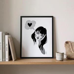 May include: Black and white framed art print featuring a pensive woman with black hair, resting her head on her hand. A thought bubble with a black heart floats above her head. The artwork is displayed on a wooden shelf with books and a mug.