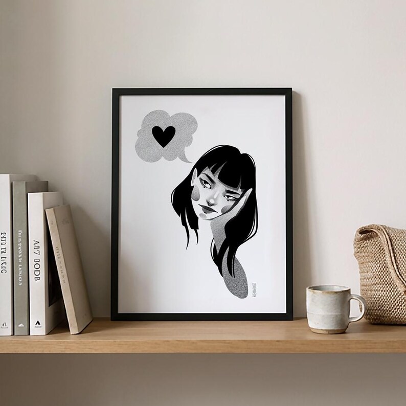 May include: Black and white framed art print featuring a pensive woman with black hair, resting her head on her hand. A thought bubble with a black heart floats above her head. The artwork is displayed on a wooden shelf with books and a mug.