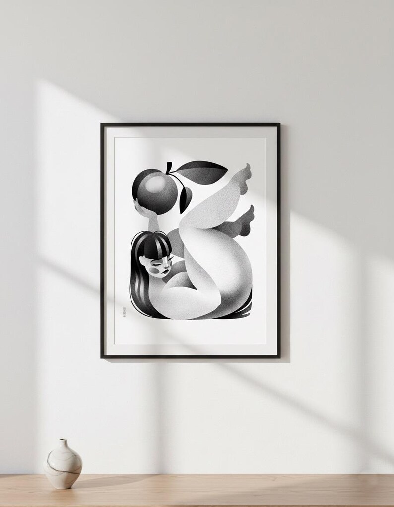 May include: Black and white art print in a black frame. The artwork depicts a stylized figure holding a fruit with a leaf. The figure has long, dark hair and is reclining. The print is mounted on a white wall.