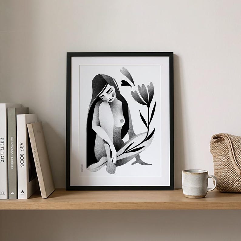 Może przedstawiać: Czarno-biały wydruk artystyczny w czarnej ramie. Grafika przedstawia siedzącą postać z długimi włosami i kwiatem. Wydruk jest eksponowany na drewnianej p&oacute;łce z książkami, kubkiem i plecioną torbą.