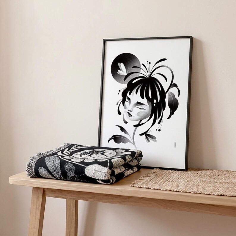 K&ouml;nnte beinhalten: Eine gefaltete schwarz-wei&szlig;e Decke mit floralem Muster und Fransen liegt auf einem Holztisch. Dahinter befindet sich ein gerahmter Schwarz-Wei&szlig;-Kunstdruck eines Frauengesichts mit floralen Elementen. Auf dem Tisch liegt auch eine kleine gewebte Matte.