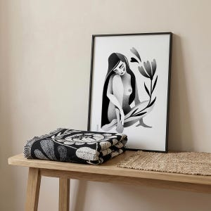 Może przedstawiać: Czarno-biały koc w kwiaty złożony na drewnianym stole. Za nim oprawiony czarno-biały nadruk siedzącej postaci z długimi włosami i kwiatem. Nadruk jest w czarnej ramie.