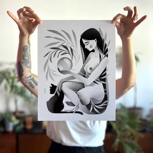 May include: Black and white art print featuring a seated figure holding a sphere, with a black cat and botanical elements. The illustration has a dot-work style, with long dark hair and a serene expression. The print is held up by a person.