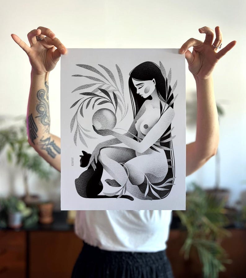 May include: Black and white art print featuring a seated figure holding a sphere, with a black cat and botanical elements. The illustration has a dot-work style, with long dark hair and a serene expression. The print is held up by a person.