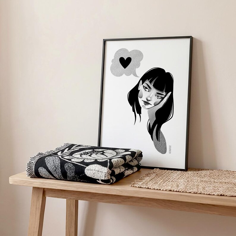 May include: A black and white framed print of a woman with a thought bubble containing a heart. A folded black and white floral patterned throw blanket rests on a wooden table. A small woven mat is also on the table.